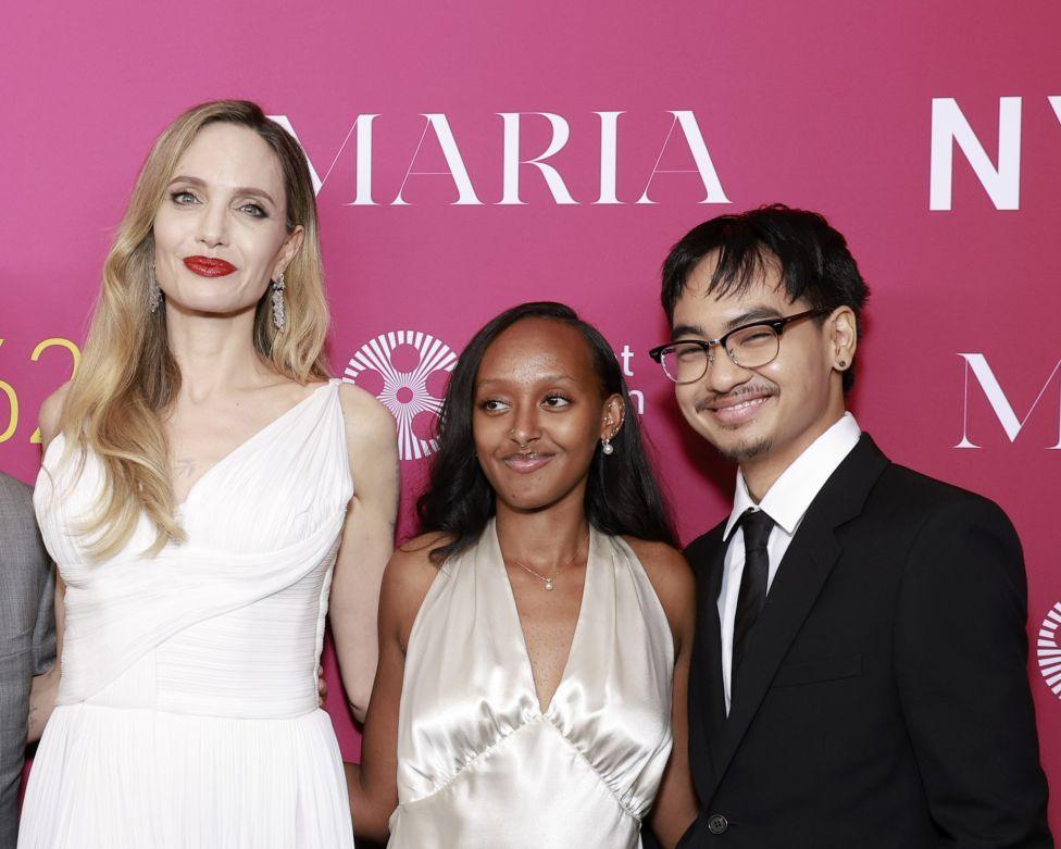 Angelina Jolie, Zahara Marley Jolie and Maddox Chivan Jolie-Pitt, in formal attire, smile at the camera in front of a pink wall 