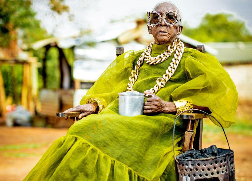 A seated Margret Chola wearing a chunky gold chain over a green taffeta like dress with puffy sleeves and a big frill for the hem holds a tin mug in her hand. Hanging off the arm of her chair is a "mbaula" filled with charcoal. She is wearing ornate framed sunglasses, the left eye frame in the shape of a D and the right frame in the shape of a C.