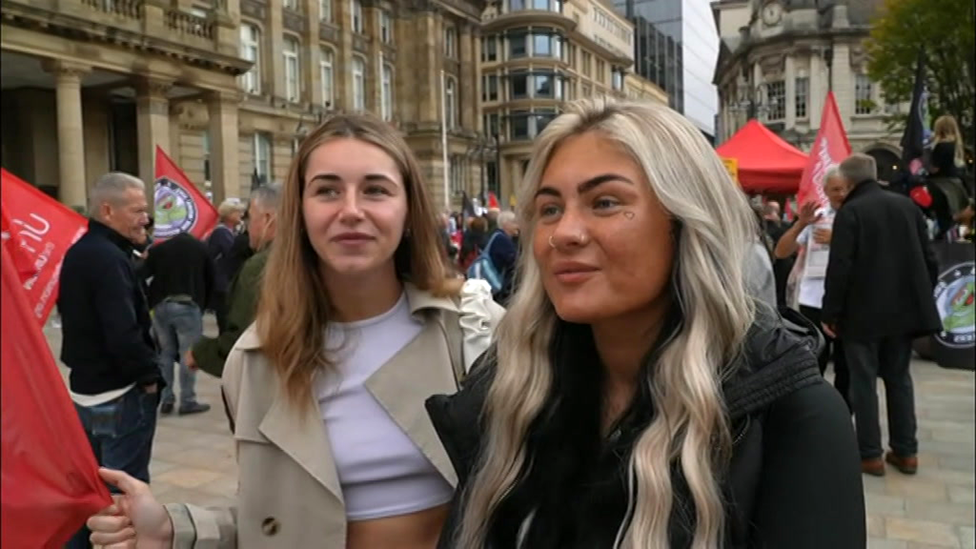 Standing next to a friend, Ronnie Mills on the right has long blonde hair and is wearing a black jacket. She is at the demo and there are people around her with red Unite flags.