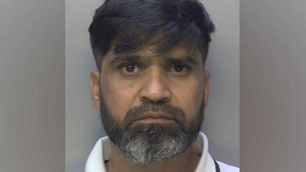 A mug shot of a man in a white polo. He has a black and white beard and black hair with a fringe.