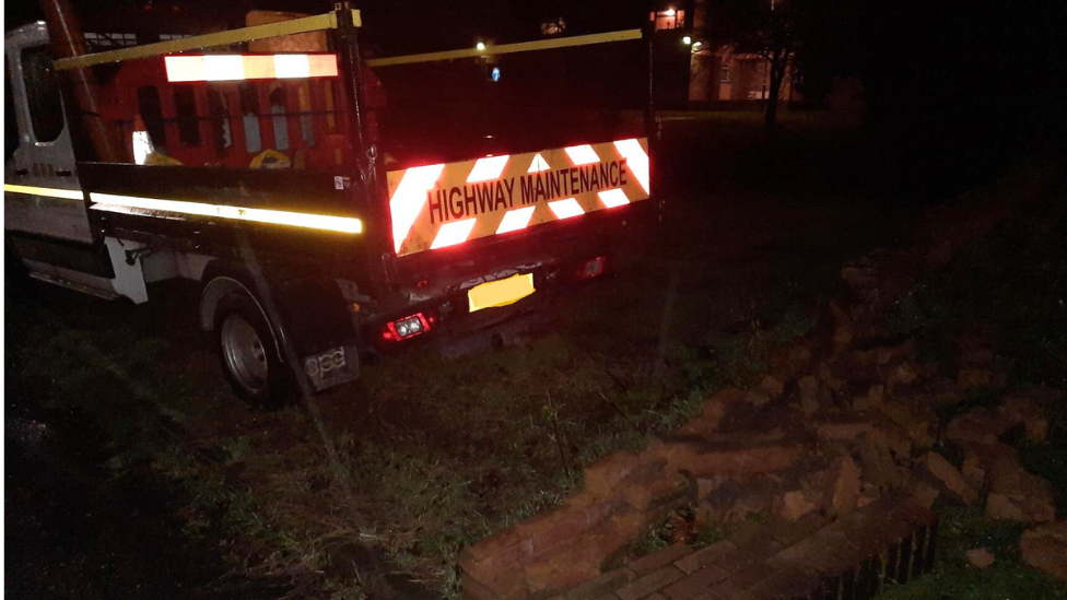 A white van marked "highways maintenance" in front of a pile of bricks. It is night-time. Some of the bricks are on the grass after the wall was hit by the van