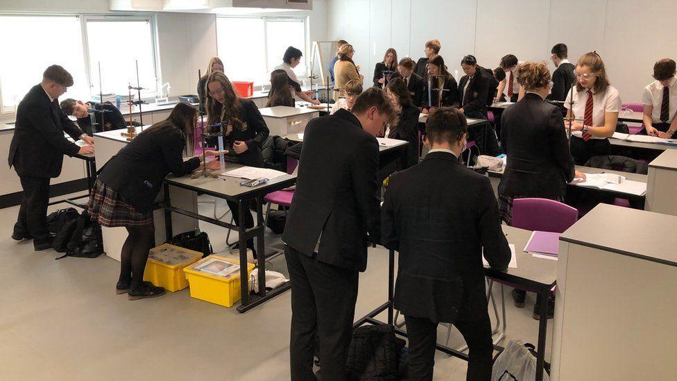 A temporary classroom full of students working on science experiments. They are all in black uniforms and learning over desks, wearing safety goggles. The classroom is a modular, plain building with no colour or decoration on the walls