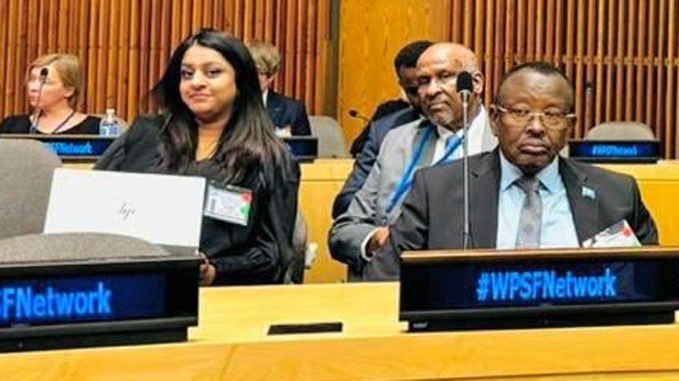 Somalia’s Family Minister Gen Bashir Mohamed Jama pictured in a conference room with other delegates, including Somali politician Abdullahi Godah Barre who is looking over his shoulder into the camera. A female delegate with long hair is seated next to them both and is also looking into the camera - New York, US - 22 October 2024