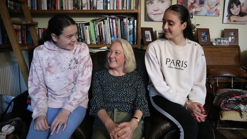 Charlotte and Georgie sit on the arms of a leather chair that their mum, Sarah, is sitting in, at their family home. They are all smiling.