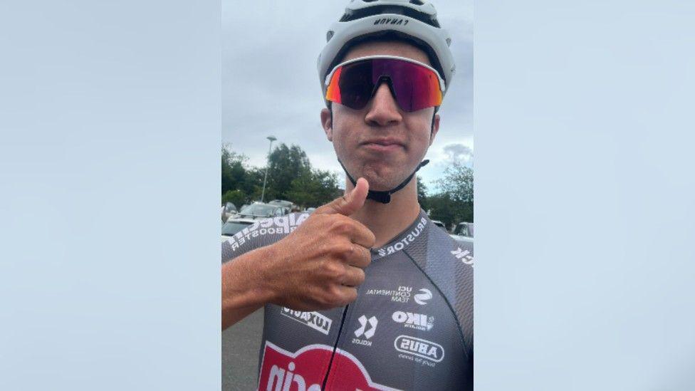 Jente Michels giving a thumbs up to the camera ahead of the Tour of the Reservoir race on 22 June. He is wearing grey cycling gear, a white helmet and red cycling glasses. Several parked cars are visible in the background.