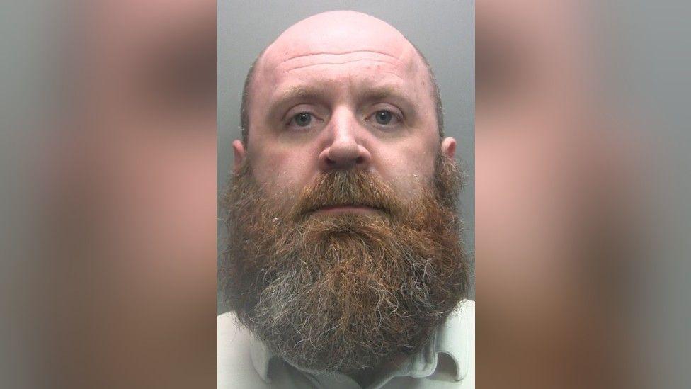 Police custody image of Kevin Clarry. He is bald with a thick ginger-and-grey beard. He is wearing a light top and looking directly into the camera.