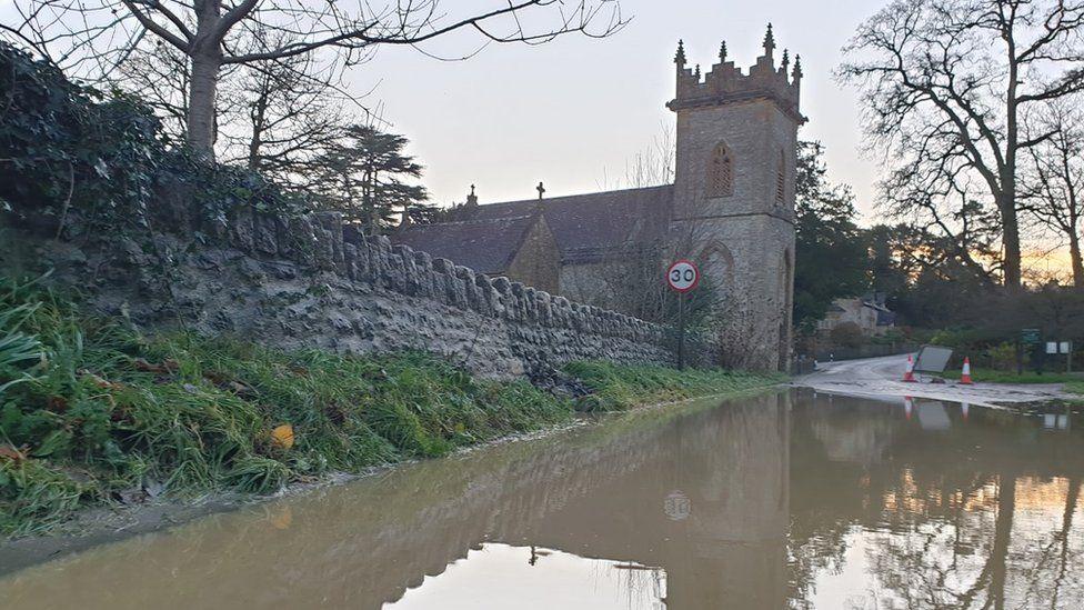 Stone wall leading from church beside road submerged in brown floodwater.