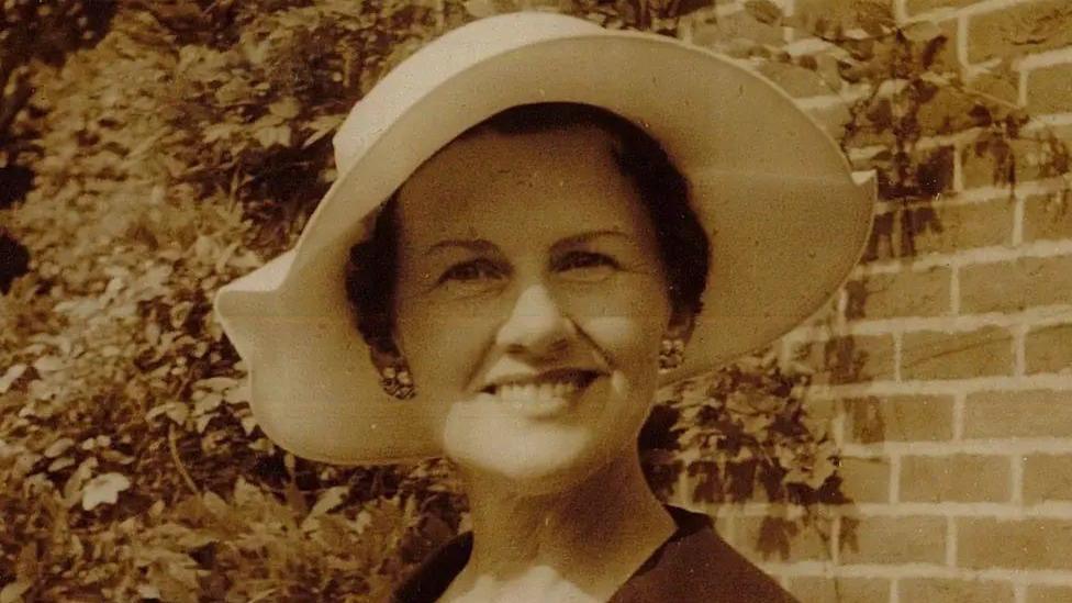 Muriel McKay photographed in a black and white sepia picture. She is wearing a cream summer hat and a dark jacket open at the neck, and cluster earrings. She is smiling at the camera and standing in front of a brick wall with a trailing plant like wisteria growing in the background.