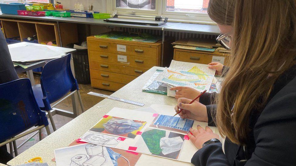 A girl draws on some paper during an art class at Plume Academy. Only the side of her head can be seen, but she is wearing glasses and has long brown hair. The paper is on a table with other sheets of paper. In front of the girl are chairs and another table. At the back of the picture is a worktop with various classroom tools, such as scissors and pens, on it.