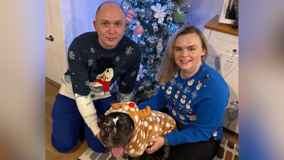 Couple James and Emily Wathey kneeling in front of a Christmas tree with their pet Staffordshire Bull terrier in between them, wearing a reindeer dog jumper