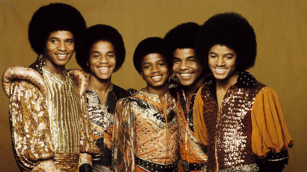 The Jackson 5 stood against a yellow background. The five members are all smiling. 