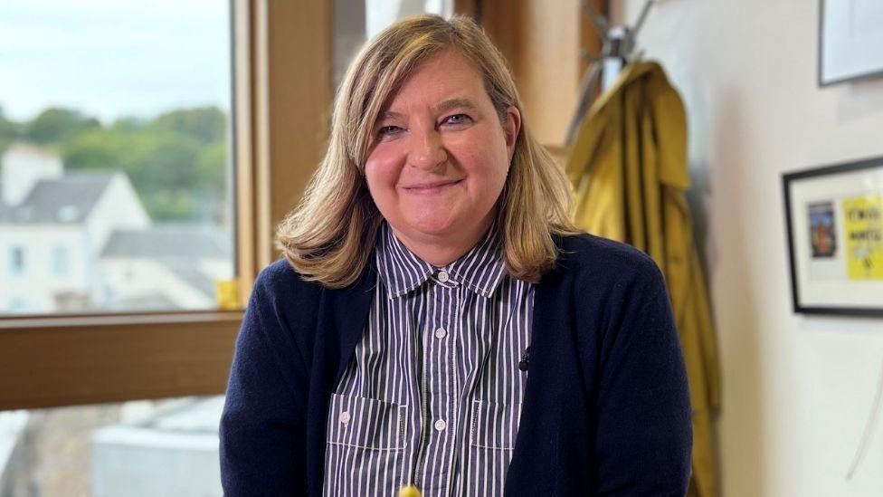 Jenni Minto wearing a dark cardigan over a striped button-up shirt, standing indoors near a window with a view of buildings and greenery outside; a yellow coat is hanging on the wall in the background, and framed artwork is partially visible.