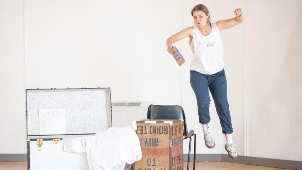 Alison Campbell is jumping into the air during rehearsal of Jane Eyre. She is wearing blue trousers and a white tank top. There are props next to her on a wooden floor.