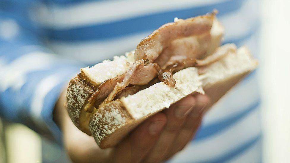 A person in a blue and white striped t-shirt holds a bacon sandwich on white bread.