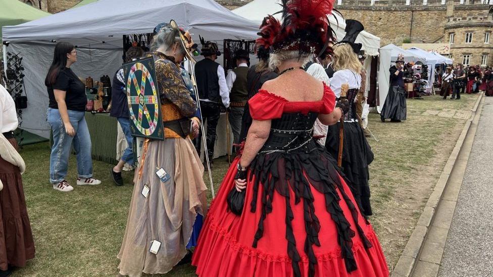 People are milling about the stalls of the festival in their outfits. One person is dressed in a flowing red and black dress, while another person is stood next to them with a long brown skirt and a circle pin board on their back. 