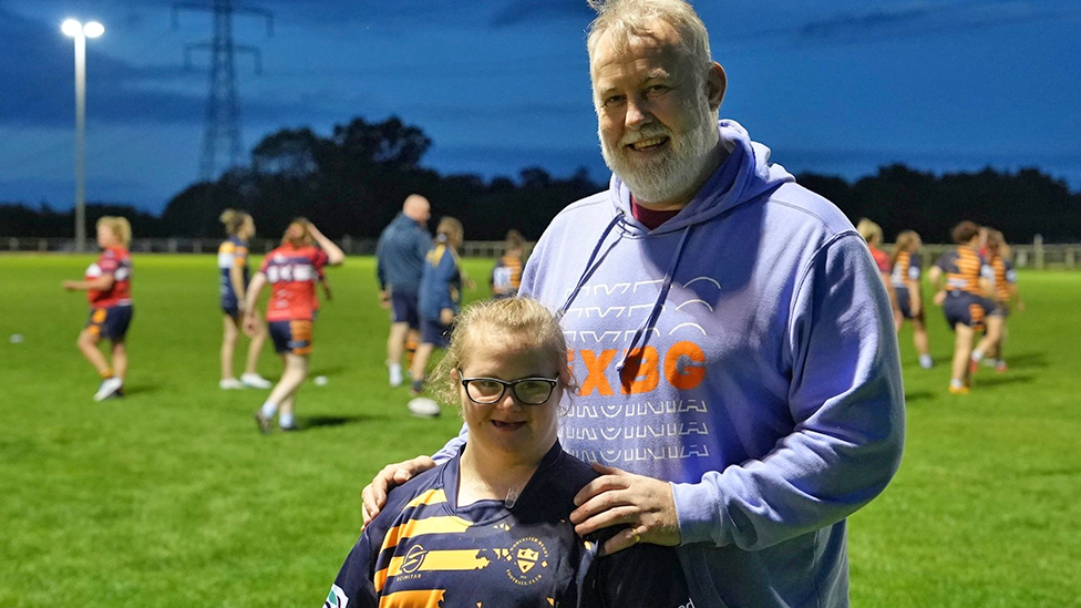 Elsie-May is wearing a Worcester shirt and smiling.  She's standing with her dad, David, on a training pitch.  Women are playing rugby in the background. 