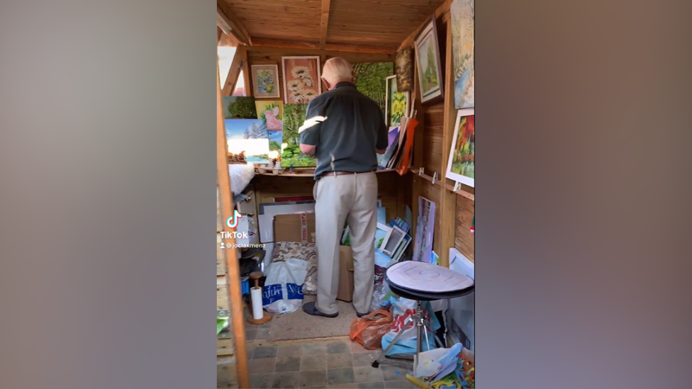 A grab of a TikTok video showing Mr Rose with his back to the camera, standing painting in his shed which is adorned with other paintings.