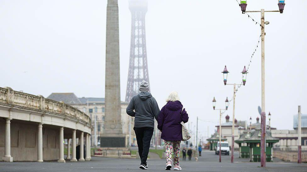 The backs of a man and woman walking down Blackpool Promenade in a misty day with a dull grey sky and fog obscuring Blackpool Tower