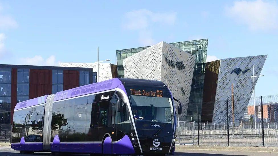 Purple bus pictured in front of Belfast's Titanic Museum, a large silver, angular building with blue sky visible 