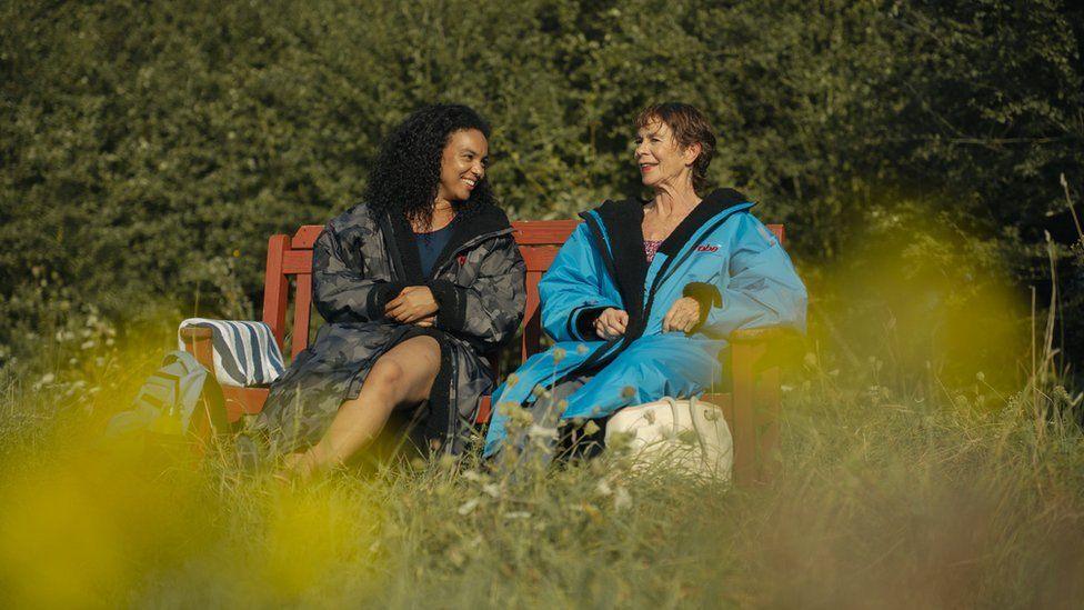 Cat White with long dark hair wearing a grey coat and blue top talking to Celia Imrie with short dark hair who is wearing a blue coat. They are sitting on a wooden bench on grass with bushes behind.