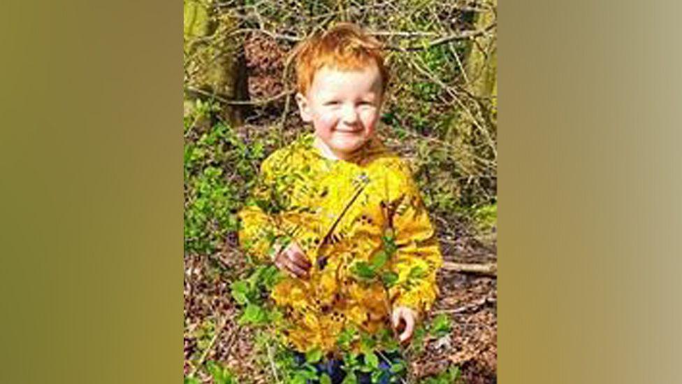 George is smiling as he hides in bushes in a park, with a tree behind him. He is wearing a yellow waterproof coat with a dinosaur design.