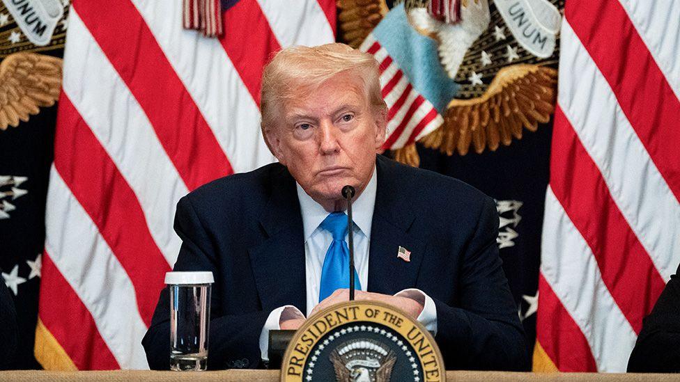 US President Donald Trump, in a dark suit, sits behind a desk at the White House with the presidential seal in front of him and a flags, including the Stars and Stripes, behind him