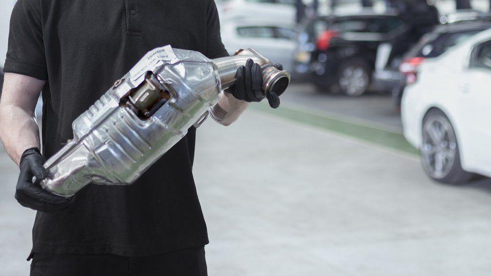 The torso of a man in black holding a silver-coloured catalytic converter