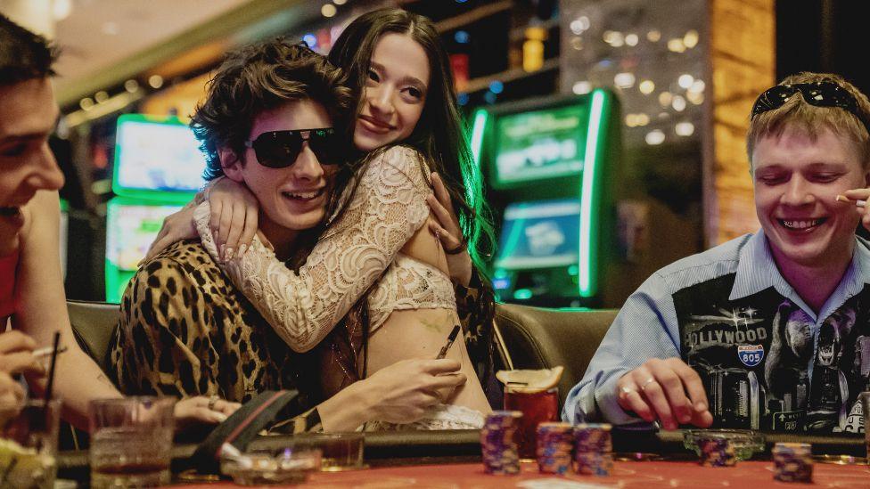 Mikey Madison and Mark Eidelstein in Anora. Mark Eidelstein's character is sitting down at a poker table and is wearing glasses and leopard skin jacket. Mikey Madison is wearing a cream coloured dress and sitting on his lap. They are both smiling. 