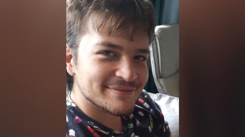 Ezra Tilford-Key has long dark hair and a beard and moustache. He is wearing a black hoodie-style top with random patterns in multiple colours. The edges of the image have been blurred for stylistic reasons to meet the picture template format.