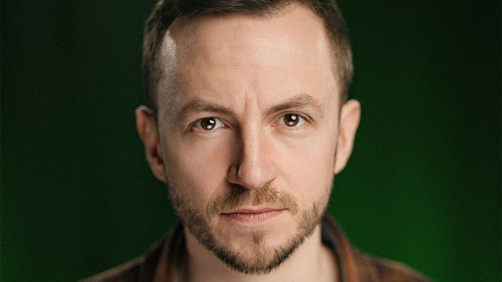 The voice over artist, Alex Jordan, is a man with a brown cwiff and a beard. He's wearing a shirt and a t-shirt underneath. On a black and green background. 