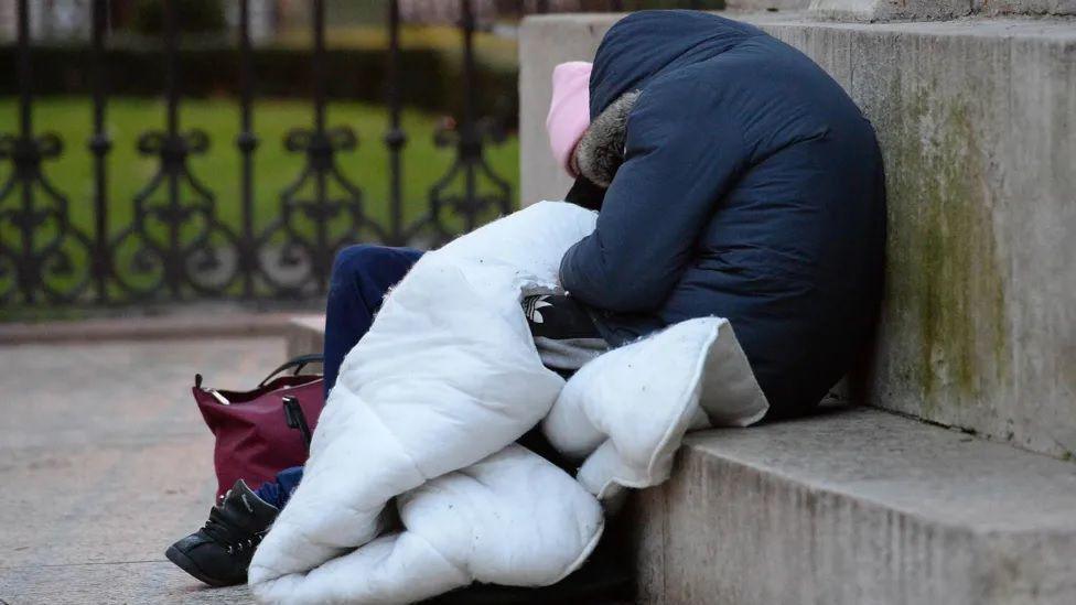 Two people sit outside on a concrete wall clutching bedding 