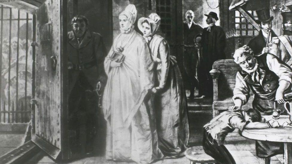 A black-and-white drawing of Elizabeth Fry in Newgate Prison in the early 19th Century. She is approaching a wooden gaol door on the right, which is being opened by a turnkey holding a large key. Through the door can be glimpsed bars and behind them faint figures of women prisoners. Behind her is another woman and behind them two men standing at a door. To the far right is a boy and man at a wooden table.