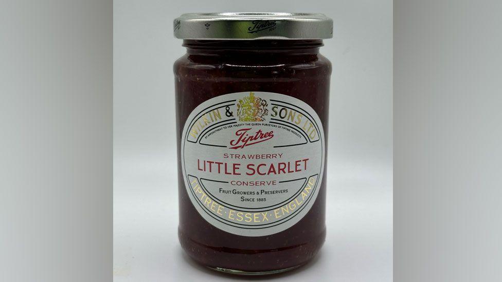 A glass jar filled with red strawberry jam. It has a white label says Tipree strawberry Little Scarlet conserve, Fruit Growers & Preserves since 1885 and Tipree Essex, England.
