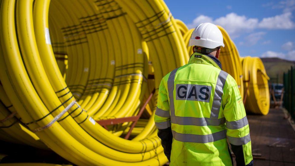 A worker stands wearing a hard hat and a high-visibility jacket with "Gas" written on the back in front of large coils of piping. The bright yellow pipes are stored outdoors