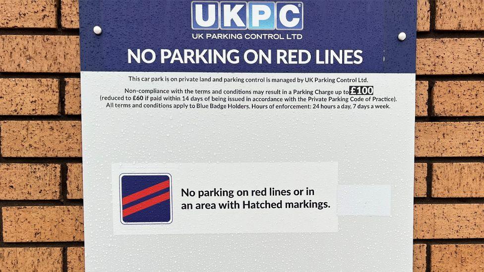 A close-up of the sign warning about fines attached to a brick wall. It says: "No parking on red lines" in capital letters, with warnings of a £100 fine for "non-compliance".