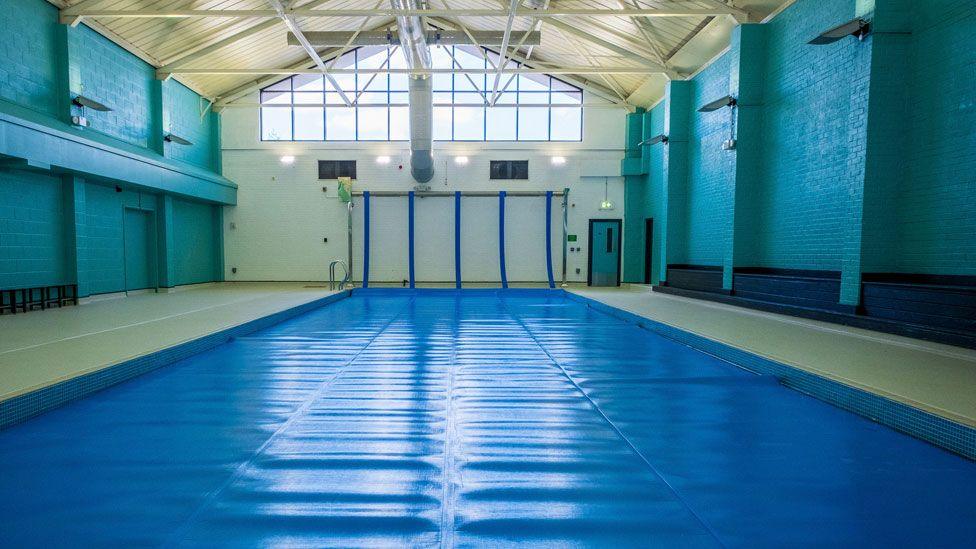 A refurbished inside swimming pool in a large hall. The water is covered with a blue cover, its brick walls are painted in turquoise and navy and its roof is painted cream. There is a large window in the far wall.