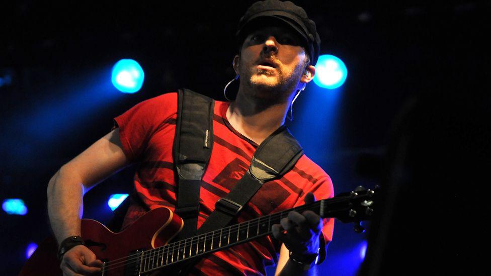 Guitarist Jez Williams, wearing a black peaked hat and a red T-shirt, plays a guitar in front of bright blue lights