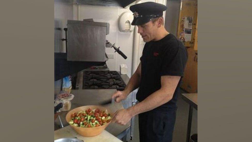 George Rudd in a blue firefighter's cap, t-shirt and trousers, preparing a salad in a kitchen