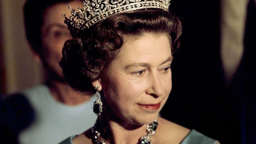 Close-up head and shoulders portrait of Queen Elizabeth II in 1975; she is looking down away from the camera while wearing a crown and an emerald necklace