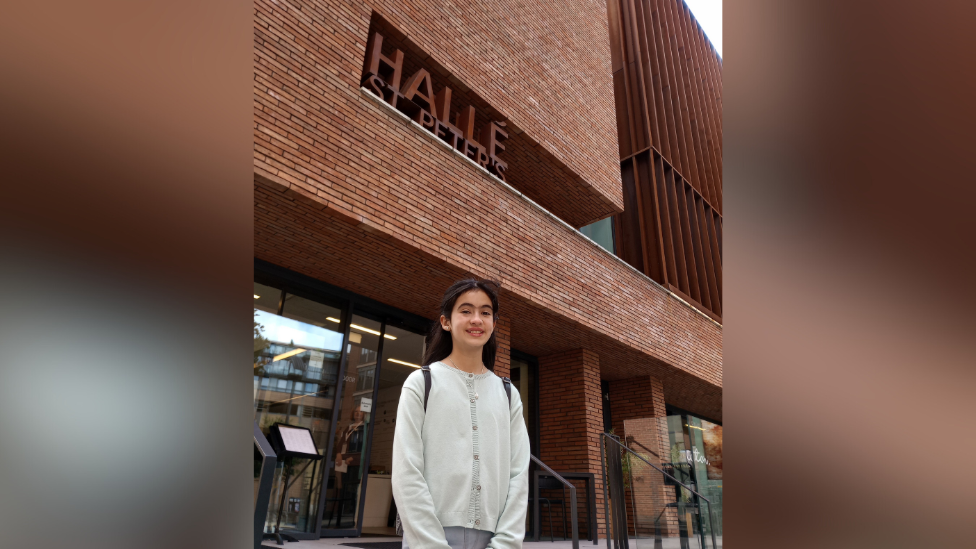 Zoe standing outside the Hallé St Peter's in Manchester. She has long dark hair partially tied back, wearing a grey buttoned up cardigan and a backpack. The building is modern with brown bricks and glass doors at the bottom.