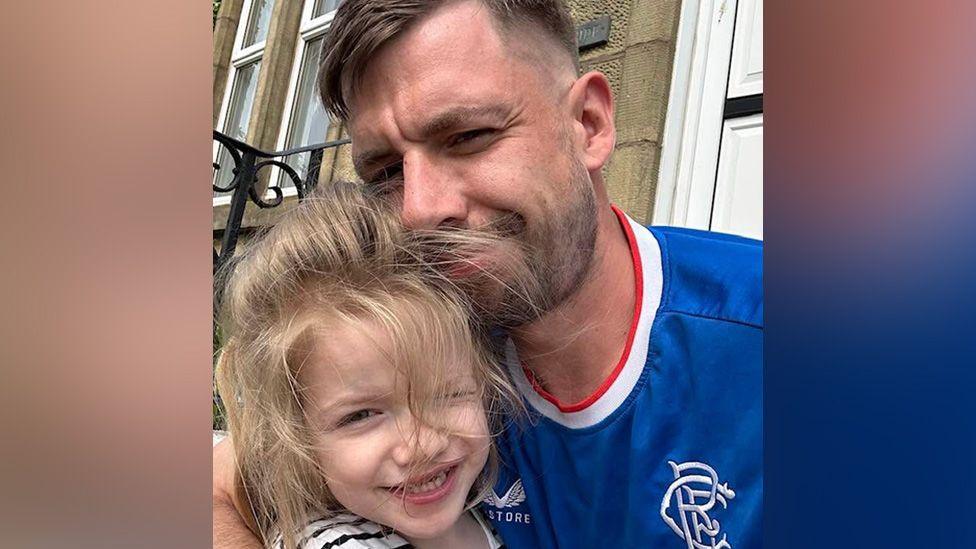 A man in a blue Rangers home top has his arm around a little girl with blonde hair. They are both smiling for the picture and appear to be standing outside a property.