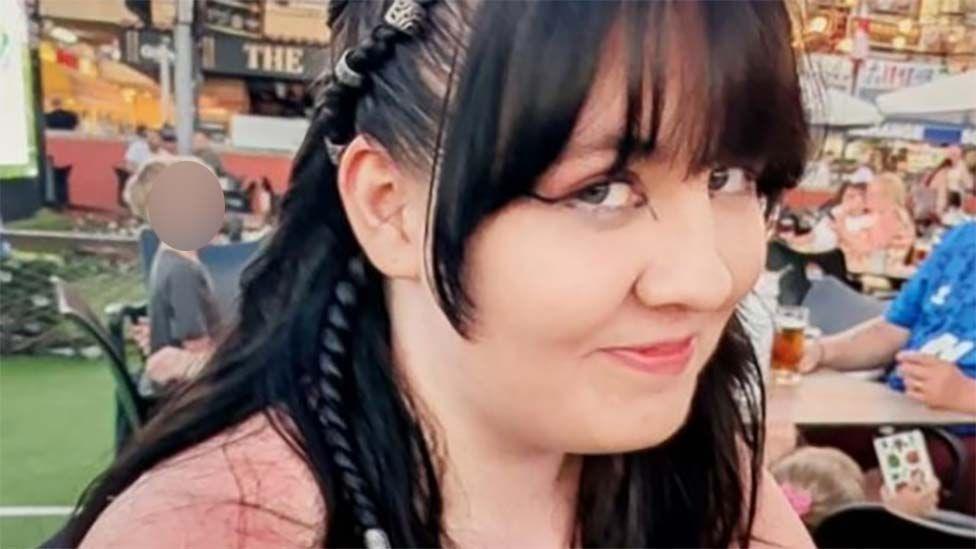 A teenage girl with long dark hair with small braids in. She is sat in a busy outdoor restaurant and is smiling and looking at the camera.