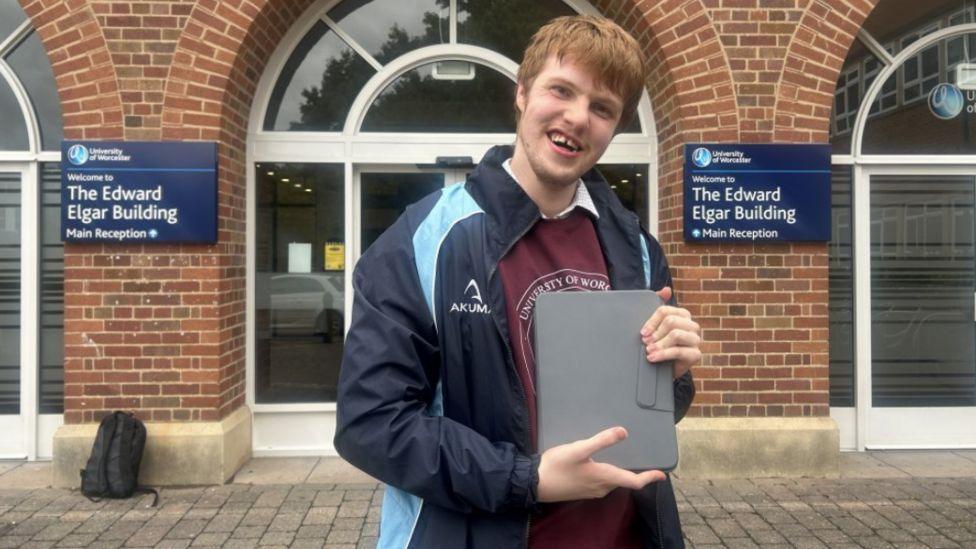 Man smiling while holding a grey tablet outside a red brick building 