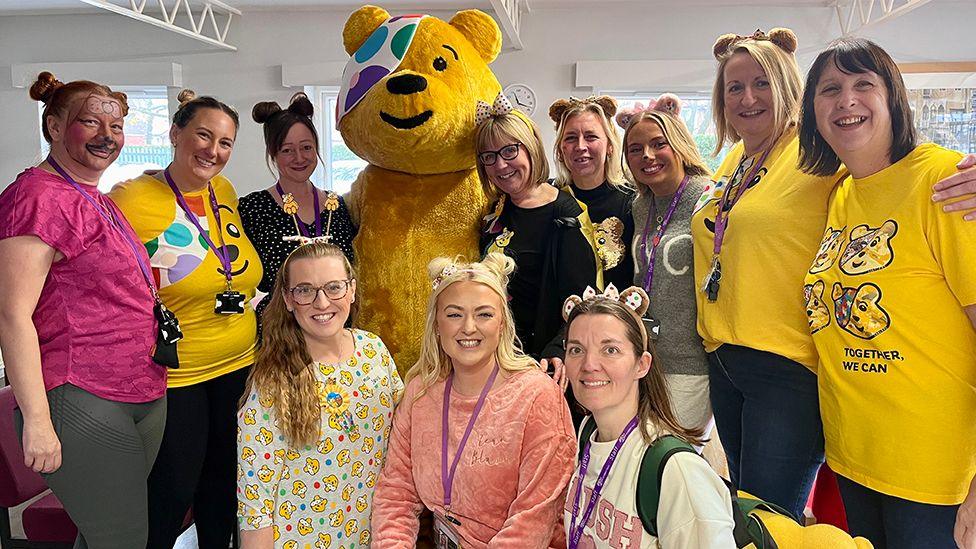 Staff at All Saints’ Church of England Infant & Junior Academy in Hessle wearing Pudsey ears and yellow Pudsey T-shirts crouch and stand around Pudsey 