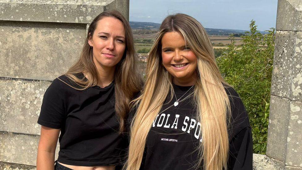 Nina Wilson and Olivia Cusack stand shoulder-to-shoulder in front of a wall overlooking a countryside valley. Nina has long, brown hair and wears a black, cropped t-shirt. Olivia has long, blonde hair and wears a black pullover hoodie with a silver heart locket around her neck.