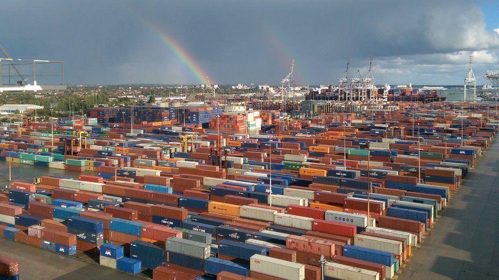 Lines of containers, white, blue and rust-coloured with grey coudy sky and rainbow in the distance