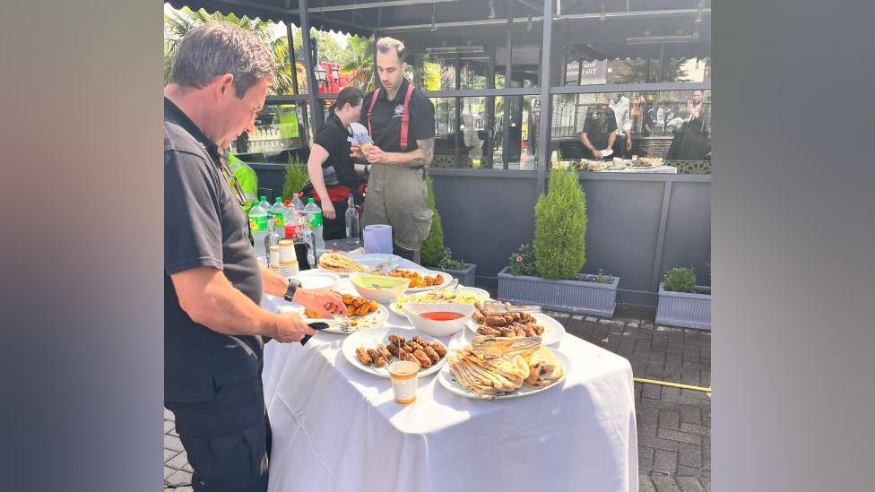 A man in a black t-shirt and trousers is standing next to a table full of food and drinks. A firefighter is holding a cup and standing on the opposite side of the table