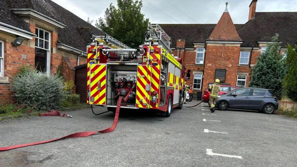 Fire engine with house coming out of rear alongside two-storey brick library building.  Two firefighters are visible.