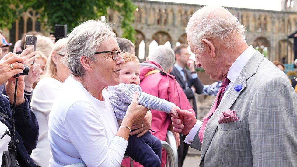 King Charles shakes the hands of a little child with red hair being held up by a woman with grey hair and glasses in a lilac jumper