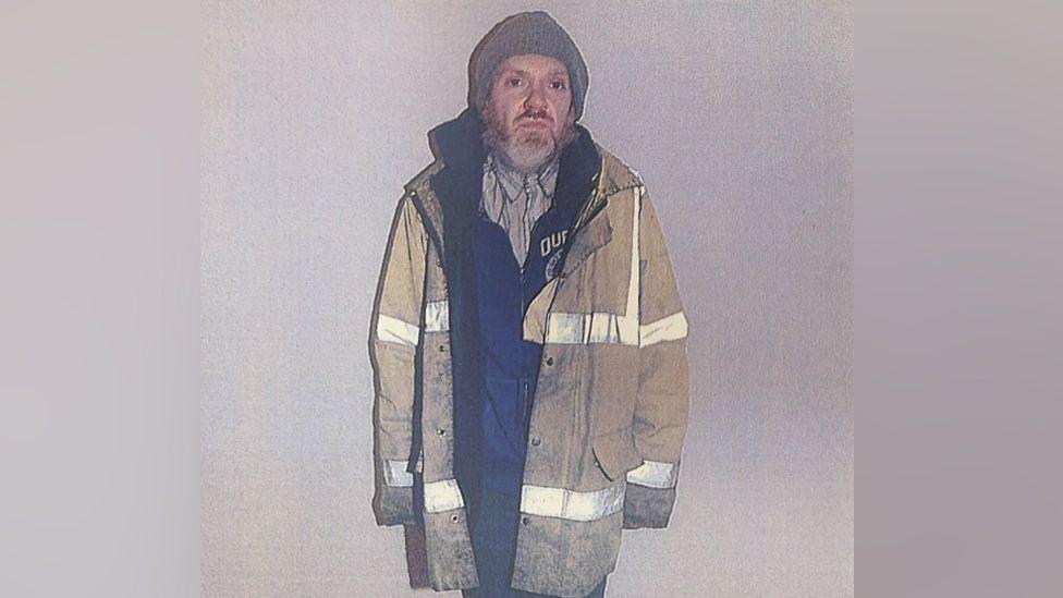 Roy Barclay in a yellow hi-vis jacket that finishes mid-thigh. He is also wearing a grey beanie, a stripy shirt, and a dark fleece zipped up